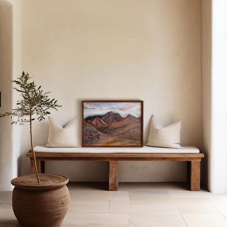 May include: A wooden bench with two cushions and a framed painting of a mountain landscape with pink and purple hues. A small olive tree in a woven basket sits on the left side of the bench.