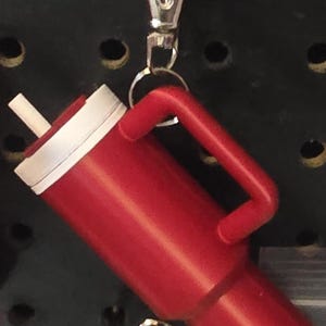 May include: A red tumbler with a white rim and a handle, featuring a white straw. The tumbler is attached to a silver clip. The background is a black pegboard with several holes.