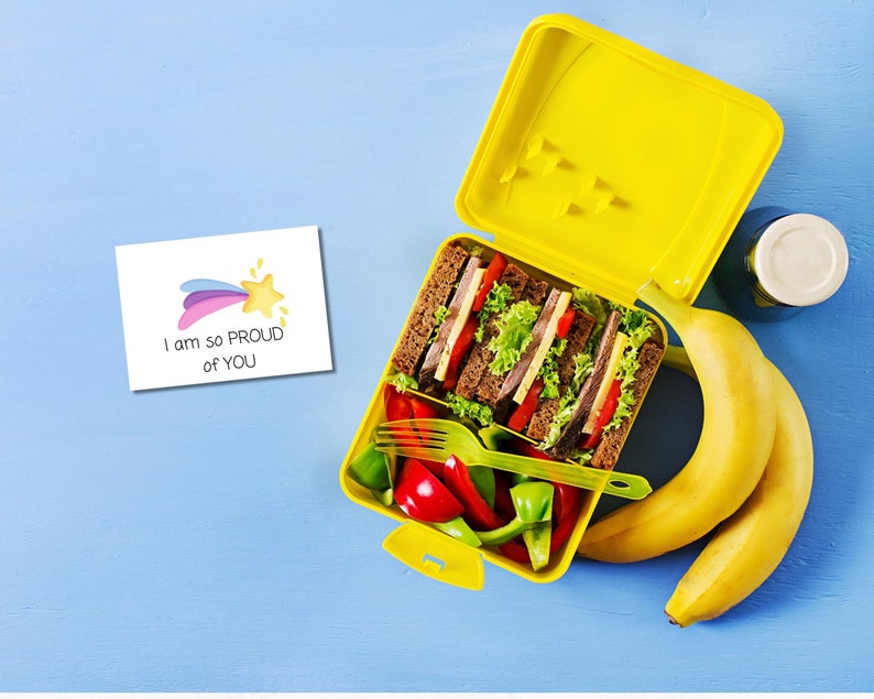 May include: A yellow lunchbox with a sandwich, vegetables, and bananas inside. A note with the text "I am so proud of you" and a rainbow star is on the blue background.