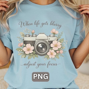 May include: Light blue t-shirt featuring a vintage camera design with pink flowers and the text "When life gets blurry, adjust your focus." The image includes a "PNG" watermark.