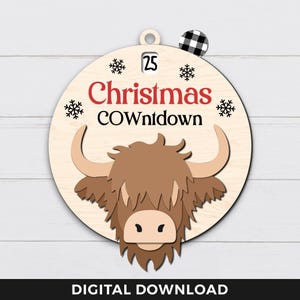 May include: A round wooden ornament featuring a brown Highland cow head illustration. The ornament has the text "25 Christmas COWntdown" printed on it.