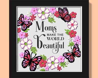 以下が含まれることがあります： 「Moms MAKE THE WORLD BEAUTIFUL」の文字が書かれた額縁入りのアートワーク。花のリースと蝶が周りを囲んでいます。花はピンク、白、赤で、緑の葉があります。蝶は黒とピンクです。フレームは黒です。