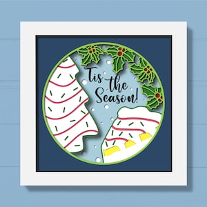 May include: A white frame with a blue background and a circle in the center. The circle has a white and red striped Christmas tree, a white and red striped gingerbread house, and green holly leaves with red berries. The text "Tis the Season!" is written in black.