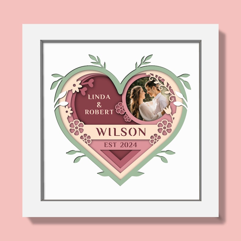 May include: A framed heart-shaped artwork with a photo of a couple. The design includes layers of pink, green, and burgundy, with the names "LINDA & ROBERT" and "WILSON EST 2024" displayed. Floral and leaf cutouts decorate the edges.