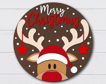 Puede incluir: Un círculo marrón con las palabras "Merry Christmas" en letras blancas y rojas. Una cara de reno de dibujos animados con un gorro de Papá Noel rojo está en el centro del círculo. El reno tiene una nariz roja y cuernos marrones con estrellas blancas colgando.