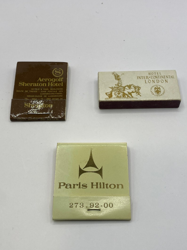 May include: Three matchbook covers. The first is brown with gold lettering and the text "Aerogolf Sheraton Hotel". The second is white with gold lettering and the text "Hotel InterContinental London". The third is white with gold lettering and the text "Paris Hilton 273.92-00".