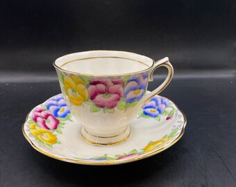 May include: A vintage teacup and saucer set with a floral pattern. The cup and saucer are white with a gold trim and feature a colorful floral design of pansies in shades of yellow, purple, blue, and green.