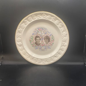 May include: A white ceramic plate with a gold rim features a commemorative image of Prince Charles and Princess Diana. The plate is decorated with a floral design and the text "Charles Diana 1981".