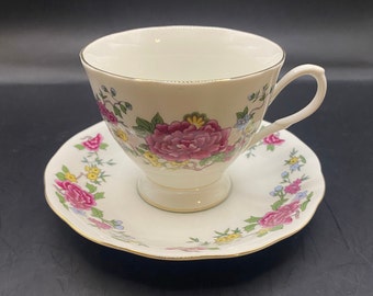 May include: A white teacup and saucer set with a floral design. The teacup features a large, pink peony and other colorful flowers. The saucer has a matching floral pattern and a gold trim. The set is on a black surface.