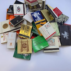 May include: A collection of vintage matchbook covers featuring various businesses and locations. The matchbooks are in various colors and designs, including red, blue, green, and yellow. Some of the matchbooks have text on them, such as "The Village Inn", "Chez Claude", and "Red Rock Village Motel".