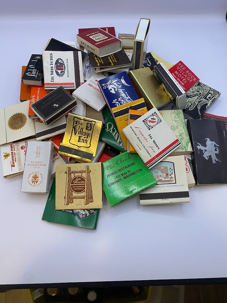 May include: A collection of vintage matchbook covers featuring various businesses and locations. The matchbooks are in various colors and designs, including red, blue, green, and yellow. Some of the matchbooks have text on them, such as "The Village Inn", "Chez Claude", and "Red Rock Village Motel".