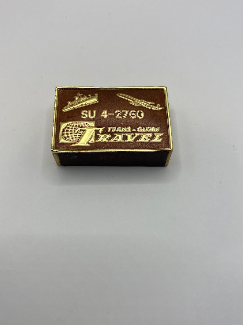 May include: A brown matchbox with gold accents featuring a globe, airplane, and ship. The text on the matchbox reads "SU 4-2760 TRANS-GLOBE TRAVEL".