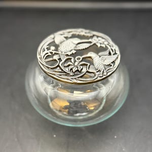 May include: A clear glass jar with a decorative silver lid. The lid features a detailed design of hummingbirds and floral elements. The jar has a rounded shape and sits on a dark surface. The lid is made of metal and has a detailed relief design.