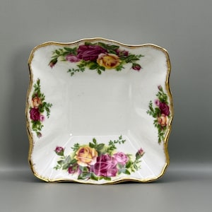 以下が含まれることがあります： 白い正方形の皿で、縁が波型で金色のトリムが付いています。皿は、黄色、ピンク、バーガンディの色合いのバラのクラスターと緑の葉で飾られています。装飾的なサービングピースです。