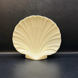 May include: A cream-colored, ceramic seashell-shaped dish. The dish has a fan-like design with ribbed detailing. The shell dish is a decorative item, suitable for holding small items or as a display piece.