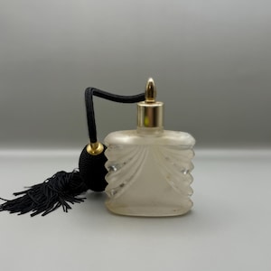 Puede incluir: Un frasco de perfume vintage con un cuerpo de vidrio esmerilado y una tapa dorada. El frasco tiene un atomizador de bulbo negro con una borla negra. El frasco es rectangular con un diseño decorativo.