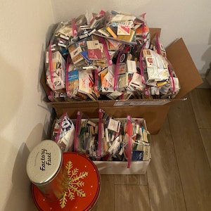 May include: A large cardboard box filled with numerous plastic bags containing matchbooks. The matchbooks are from various brands and locations. A smaller box with matchbooks is in the foreground, along with a red tin with a snowflake design and a silver tin labeled "Factory Fuel".