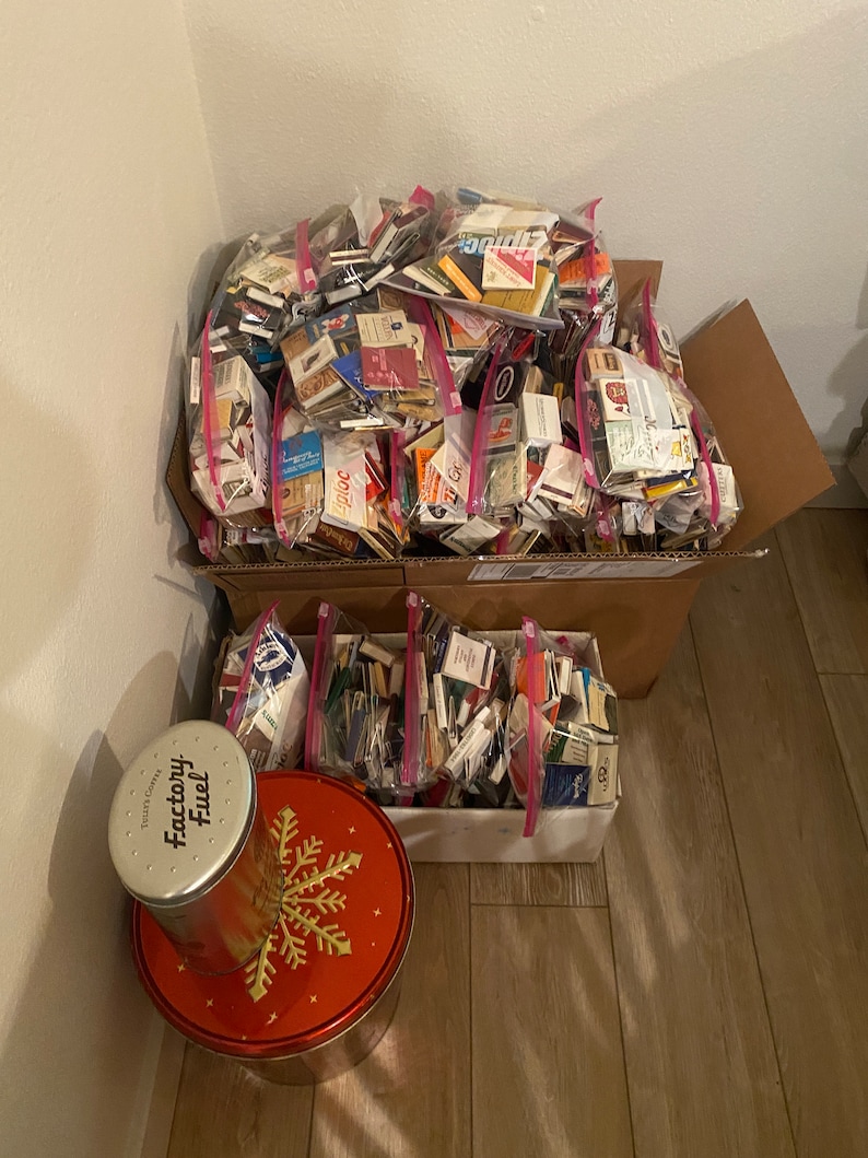 May include: A large cardboard box filled with numerous plastic bags containing matchbooks. The matchbooks are from various brands and locations. A smaller box with matchbooks is in the foreground, along with a red tin with a snowflake design and a silver tin labeled "Factory Fuel".