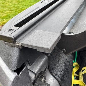 May include: A close-up shot of a black truck bed cover, with a textured surface and hinge detail. The cover is partially open, exposing the truck bed interior. Green grass is visible in the background.