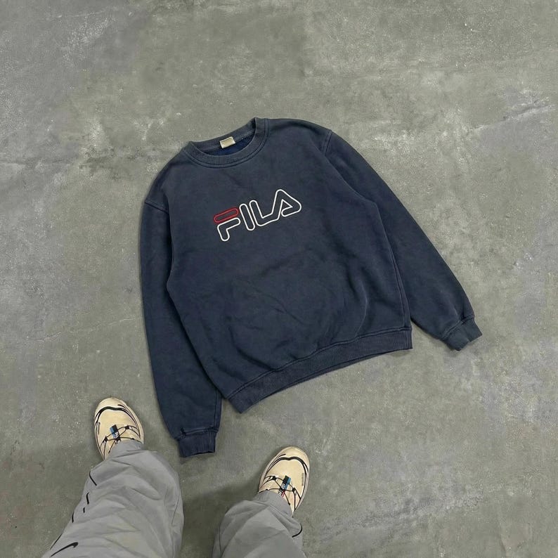 K&ouml;nnte beinhalten: Ein marineblaues Fila-Sweatshirt mit dem Markenlogo in Wei&szlig;, Rot und Marineblau. Das Sweatshirt hat einen Rundhalsausschnitt und scheint aus einem weichen Material zu bestehen. Das Sweatshirt liegt auf einer grauen Oberfl&auml;che.