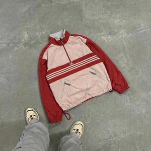 Puede incluir: Chaqueta vintage Adidas con media cremallera. La chaqueta presenta mangas rojas y un cuerpo rosa claro con una franja roja y tres rayas blancas. El logotipo de Adidas es visible. Dos bolsillos delanteros. Estilo retro.