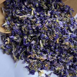 May include: A close-up of a brown paper bag filled with dried purple flower petals. The petals are scattered on a light purple surface.
