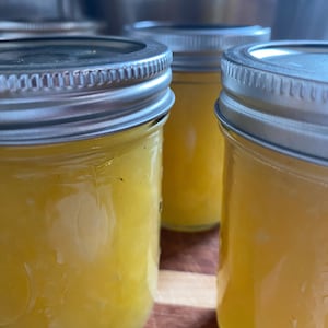 May include: Three glass jars filled with a bright yellow substance, possibly preserves or a sauce, are visible. Each jar has a silver lid. The jars are arranged on a wooden surface, with stainless steel pots in the background.