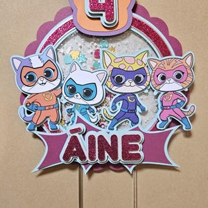 May include: A vibrant cake topper with four cartoon cat superheroes. The topper displays the number "4" and the name "AINE". The cats are dressed in superhero outfits with masks and capes, set against a glittery background.
