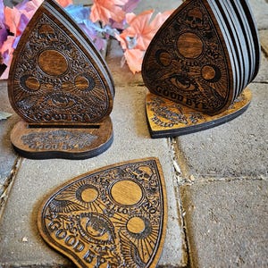 May include: A set of dark brown wooden coasters shaped like planchette pointers. Each coaster features laser-engraved details including a central eye, moon phases, a skull, and the words YES, NO, HELLO, and GOOD BYE. Two coasters stand upright in a wooden holder.