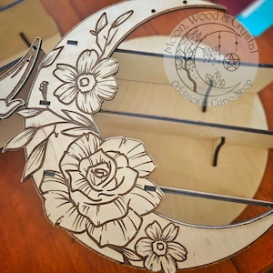 May include: A wooden crescent moon shelf with floral designs, featuring three shelves. The shelf is decorated with a rose, smaller flowers, and leaves. The text "Moon, Wood & Crystal Oddities & Curio Shop" is printed on the shelf.