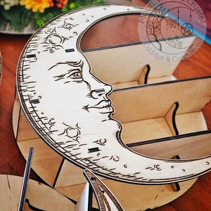 Puede incluir: Un estante de madera en forma de media luna con una cara tallada en la luna. El estante tiene cuatro compartimentos y está decorado con un diseño de estrella y media luna. El estante está etiquetado como "Moon, Wood & Crystal Oddities & Curio Shop".