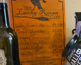 Puede incluir: Un letrero de madera naranja para "The Lucky Raven Cafe" con una silueta de cuervo y elementos del menú. El menú incluye "Lucky Raven Latte" y "Witches Brew Wine". Se ve una taza negra con "Happy Halloween Week".