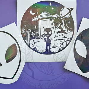 Op de afbeelding: Drie holografische stickers met alien-thema's. Eén sticker toont een alienhoofd, een andere een UFO-scène met een alien die een bloem vasthoudt. De stickers hebben een reflecterende, iriserende afwerking.