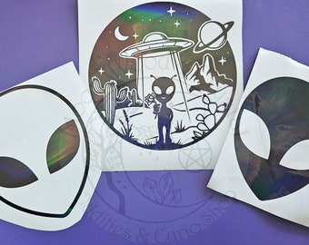 May include: Three vinyl decals featuring extraterrestrial themes. One circular decal shows an alien holding a flower under a UFO in a desert landscape with cacti and a planet. Two additional decals depict the outline of an alien head shape.