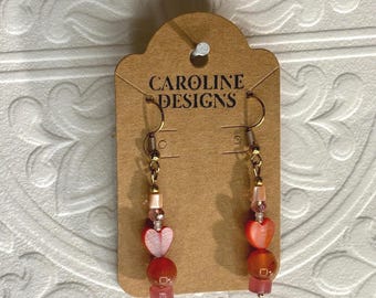 May include: A pair of dangle earrings with a heart-shaped charm and red and pink beads. The earrings are on a brown card with the text "CAROLINE DESIGNS".