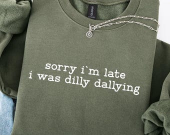 May include: An olive green sweatshirt with the text "sorry I'm late I was dilly dallying" in white. A silver necklace and a pair of blue jeans are also in the image. The sweatshirt is folded with the sleeves tucked in.