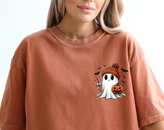 May include: A burnt orange t-shirt with a cartoon ghost wearing a beanie hat and holding a pumpkin. The ghost is surrounded by bats.