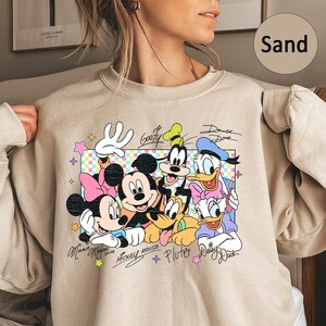 May include: Sand-colored sweatshirt featuring a colorful graphic of Disney characters including Mickey Mouse, Minnie Mouse, Goofy, Donald Duck, and Pluto. The design includes character signatures and a pastel-colored border.