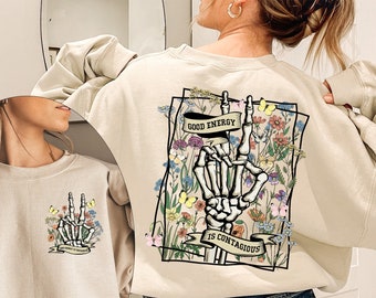 May include: A beige sweatshirt with a graphic print of a skeleton hand making a peace sign surrounded by flowers. The text "Good Energy is Contagious" is printed on a banner below the hand.