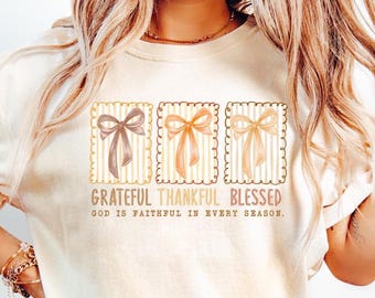 May include: Cream-colored t-shirt featuring three rectangular designs with ribbon bows in brown, peach, and tan. The text "Grateful, Thankful, Blessed" and "God is faithful in every season" are printed below the designs.