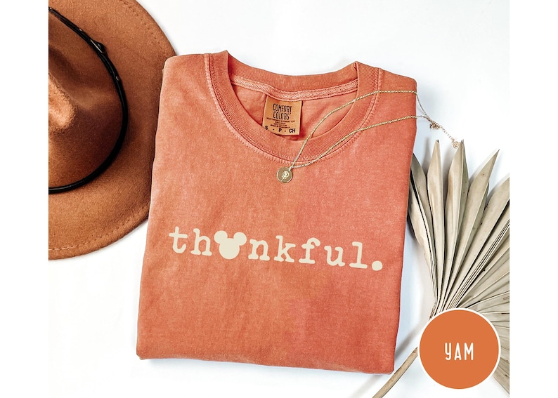 May include: A coral-colored t-shirt with the word "thankful" printed in white, featuring a Mickey Mouse head in place of the "o". A gold necklace and a brown hat are also visible.