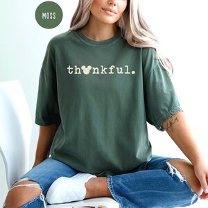 May include: A moss green t-shirt with the word "thankful" printed in white. The "o" in thankful is replaced with a Mickey Mouse head. The shirt is short-sleeved and worn with blue jeans.