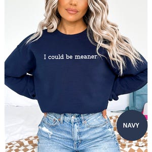 May include: Navy blue sweatshirt with the text "I could be meaner" in white. The sweatshirt is a crew neck style. The model is wearing blue jeans.