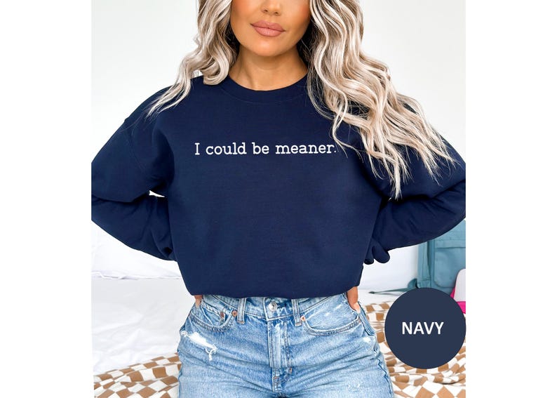 May include: Navy blue sweatshirt with the text "I could be meaner" in white. The sweatshirt is a crew neck style. The model is wearing blue jeans.