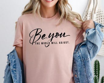 May include: A light pink t-shirt with the words "Be you, the world will adjust" in black script. The shirt is made of a soft material and has a round neckline. The model is wearing a denim jacket.