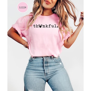 May include: A light pink t-shirt with the word "thankful" printed in black. The "o" in the word is replaced with a Minnie Mouse head. The shirt is paired with blue jeans.
