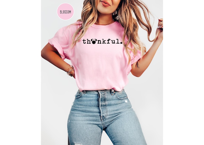 May include: A light pink t-shirt with the word "thankful" printed in black. The "o" in the word is replaced with a Minnie Mouse head. The shirt is paired with blue jeans.