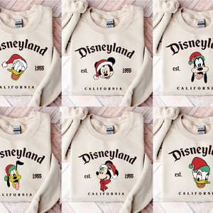 May include: Six different designs of beige sweatshirts with the text "Disneyland est. 1955 California" and a cartoon character wearing a Santa hat. The characters are Mickey Mouse, Donald Duck, and Goofy.