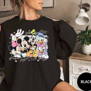 May include: Black sweatshirt featuring a colorful graphic of Disney characters Mickey Mouse, Minnie Mouse, Goofy, Donald Duck, and Pluto. The design includes character signatures and a decorative frame. The word "Black" is in a black circle.