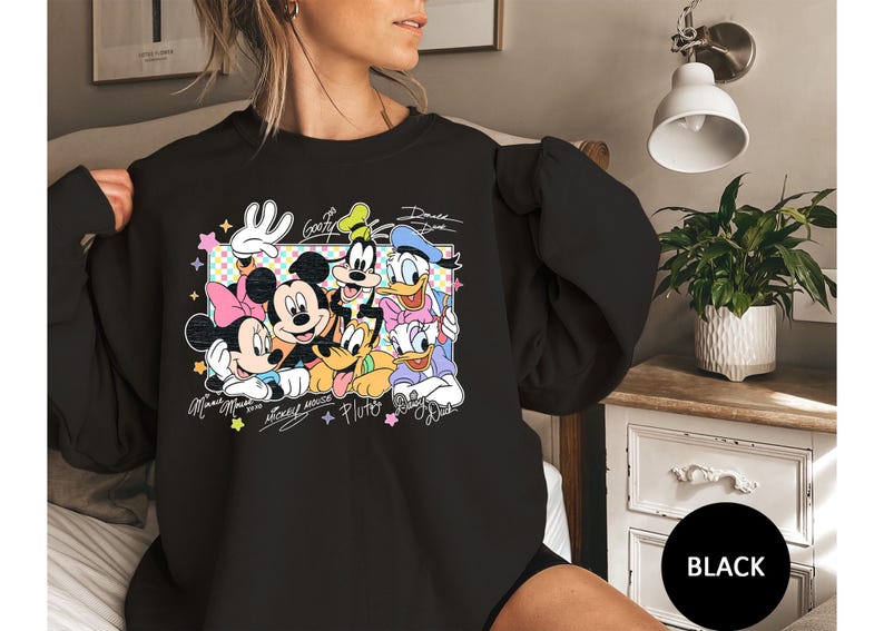 May include: Black sweatshirt featuring a colorful graphic of Disney characters Mickey Mouse, Minnie Mouse, Goofy, Donald Duck, and Pluto. The design includes character signatures and a decorative frame. The word "Black" is in a black circle.
